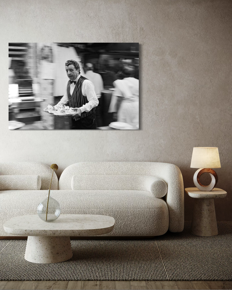 Mustavalkoinen valokuvateos canvas-kankaalla, jossa tarjoilija liikkuu ravintolassa tarjotin kädessään, tausta on sumea ja tunnelma vauhdikas.